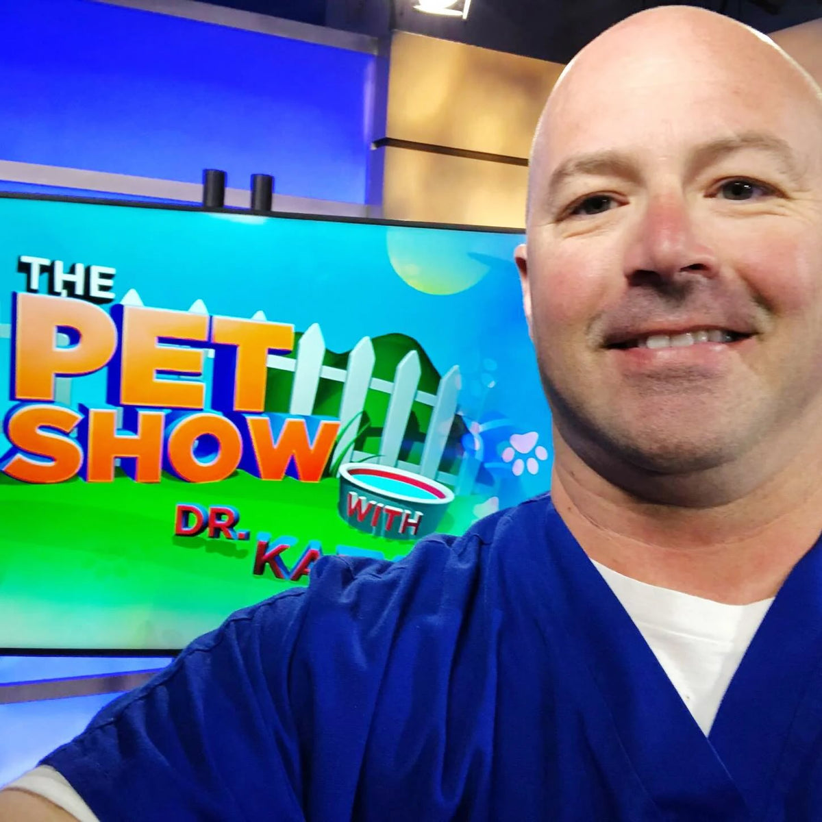 Ben Blecha in blue medical scrubs takes a selfie in front of a television screen displaying 'The Pet Show with Dr. Ka.