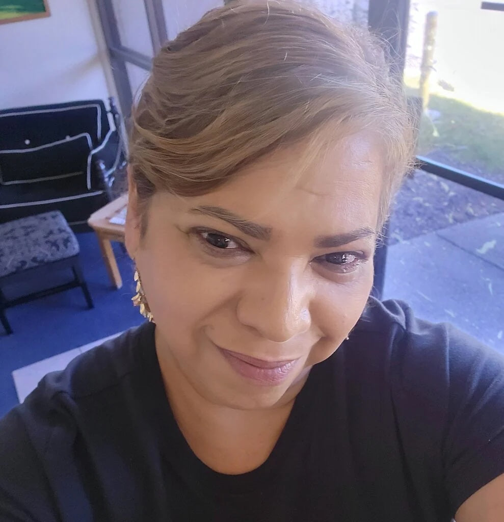 A close-up selfie of Isabel Delgado with light brown hair wearing a black t-shirt and gold earrings, looking directly at the camera in an indoor setting.