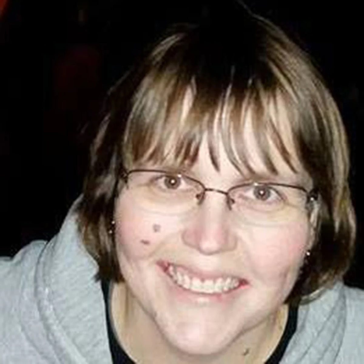 A close-up portrait of Teresa Kipp with short brown hair, bangs, and wire-rimmed glasses, wearing a light gray hoodie against a dark background.